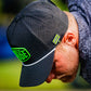 A person wearing a black perforated cap with neon green patches and a patterned shirt looks down, the brim featuring a Pins and Aces HEYDUDE - Hole 51 Ball Marker attached as a magnetic accent.