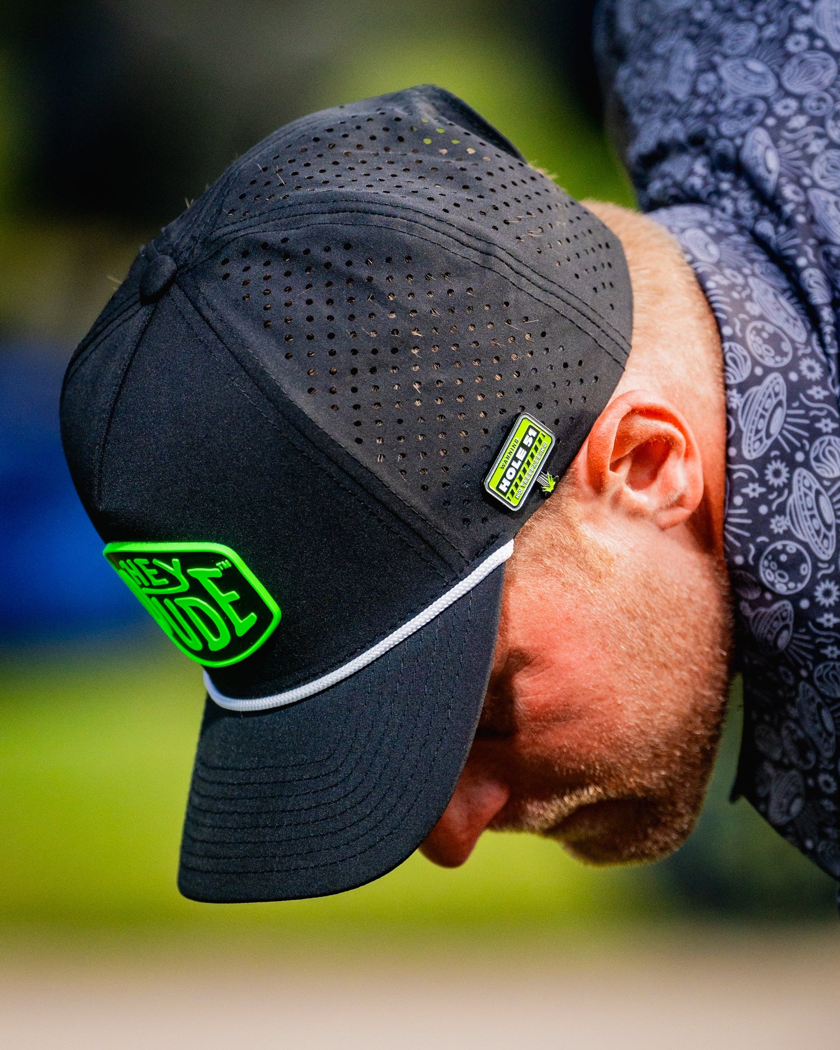 A person wearing a black perforated cap with neon green patches and a patterned shirt looks down, the brim featuring a Pins and Aces HEYDUDE - Hole 51 Ball Marker attached as a magnetic accent.