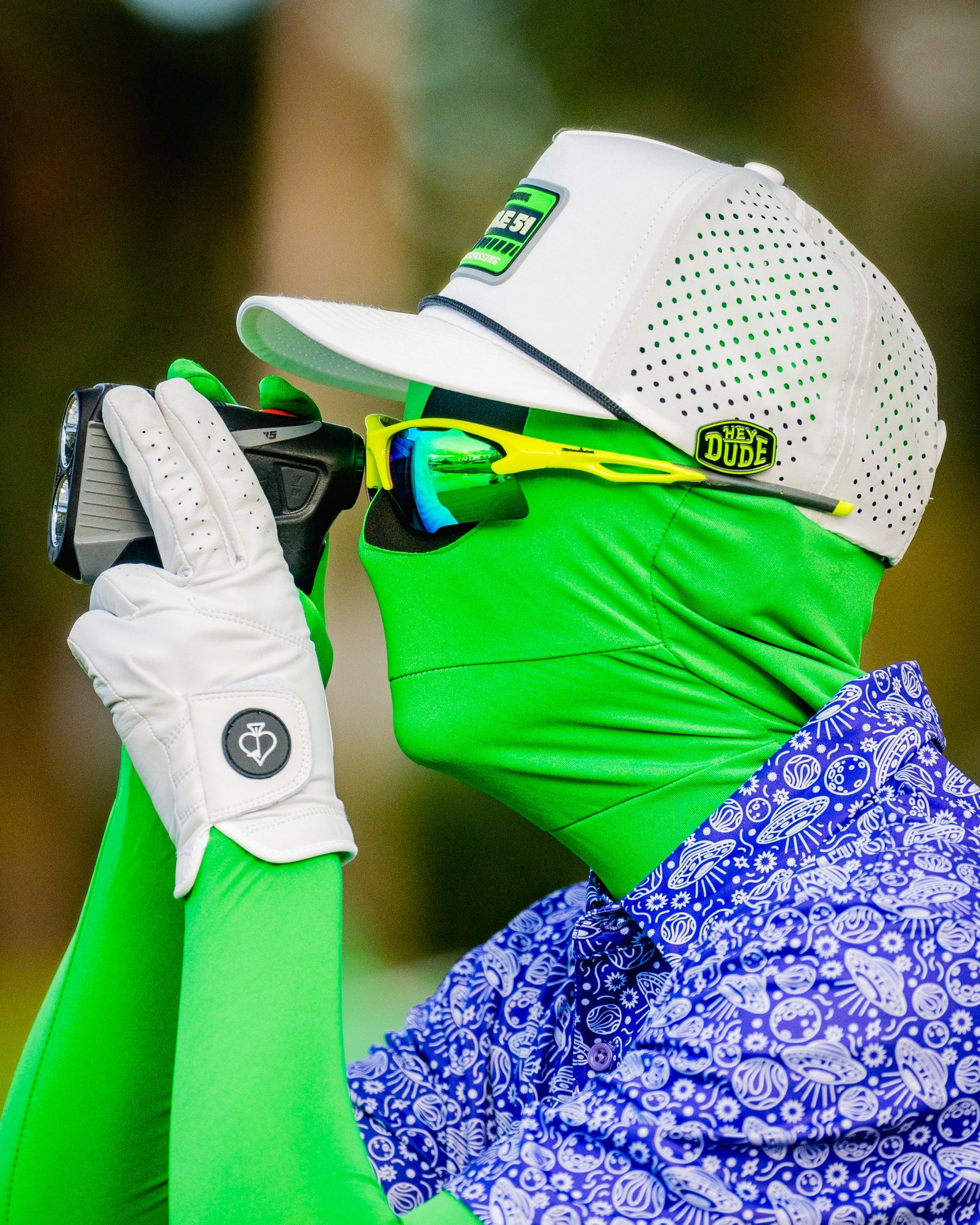 A golfer in a green suit and sunglasses, wearing a white cap and golf glove, uses a rangefinder while sporting the Pins and Aces HEYDUDE Logo Ball Marker outdoors.