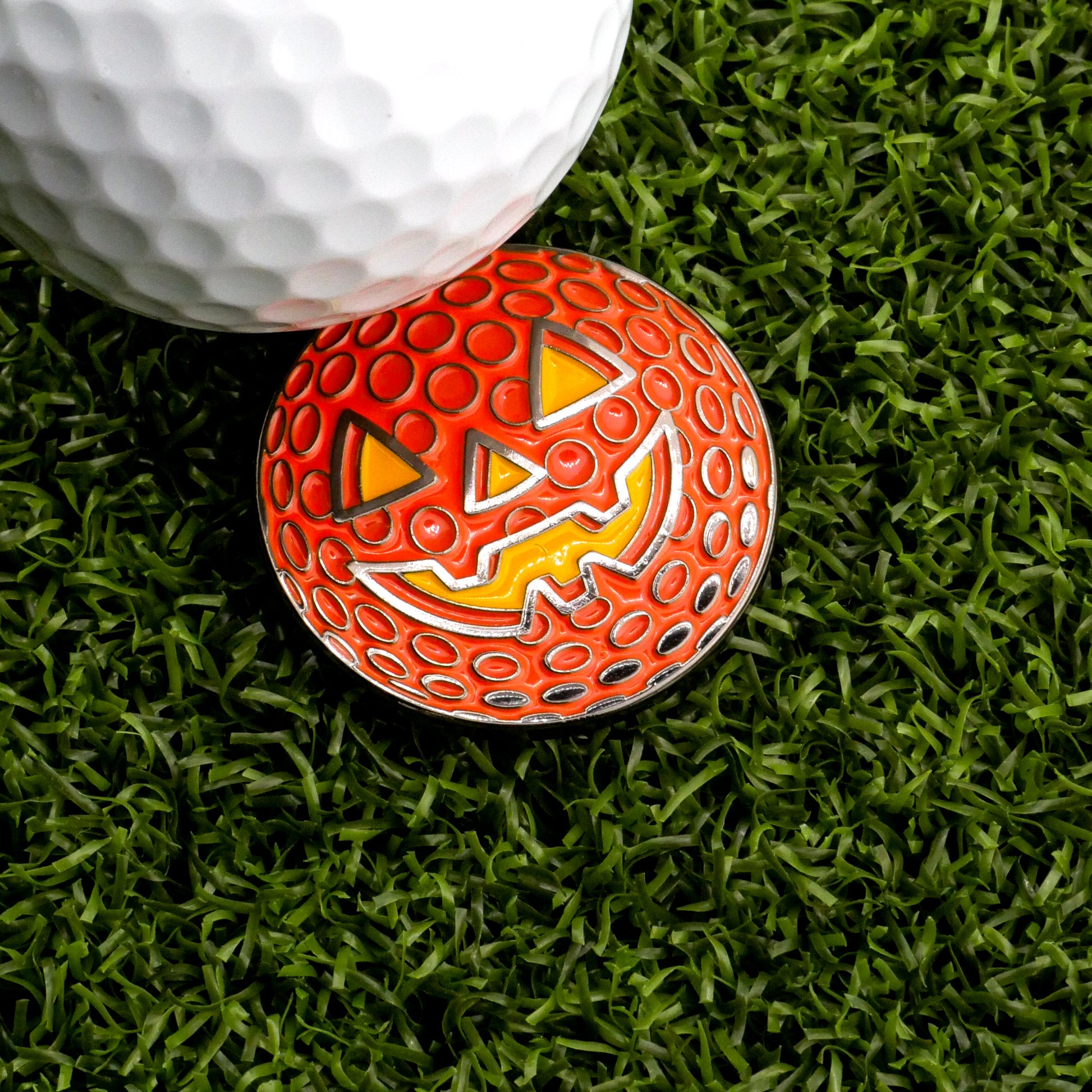 A white golf ball sits on artificial green grass next to a Ballmarkers.com Jack-o'-lantern Golf Ball Ball Marker shaped like a smiling orange pumpkin.