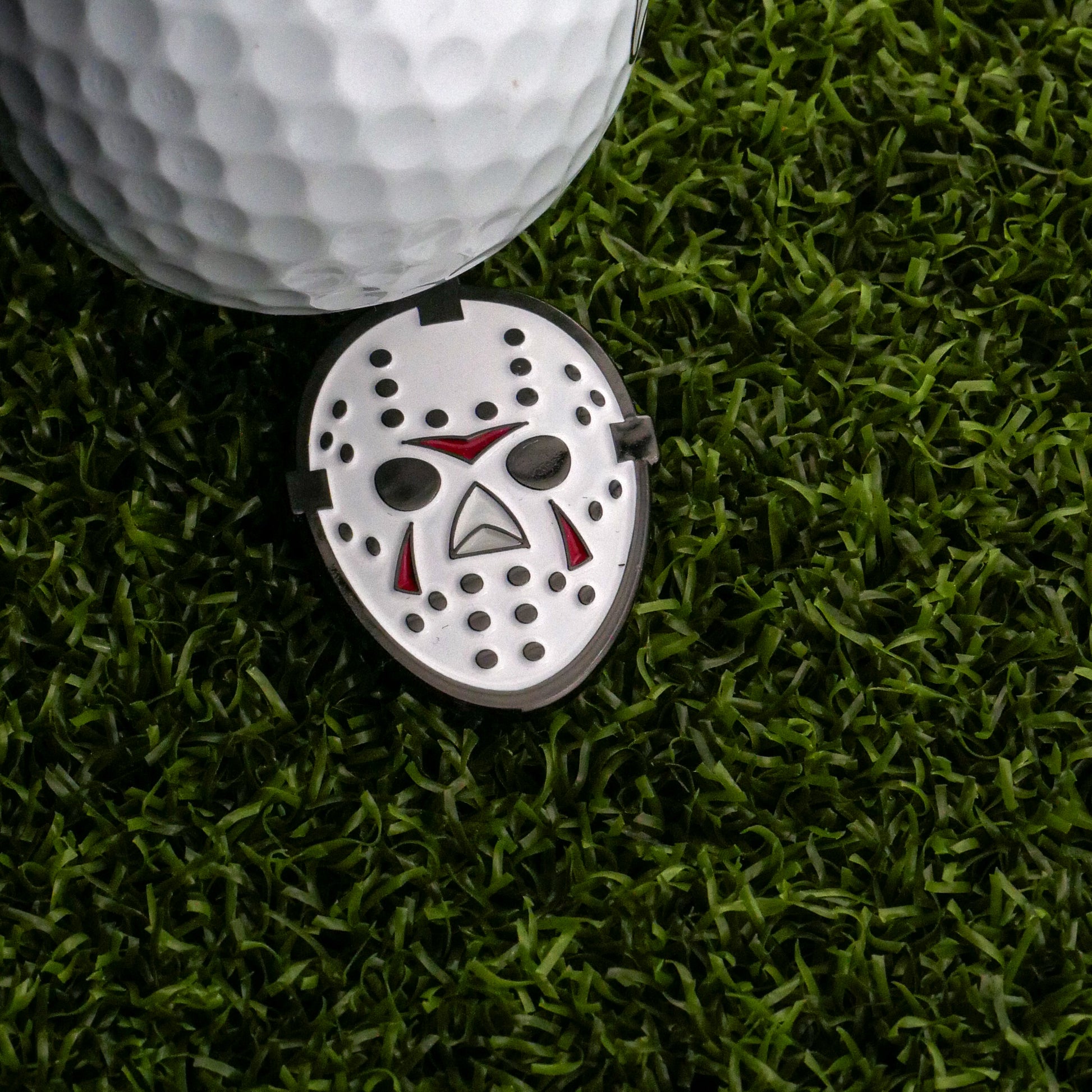 A white golf ball sits on artificial grass beside the Hockey Mask Ball Marker by Ballmarkers.com, a small metal marker with cutout eyes and red mask markings.
