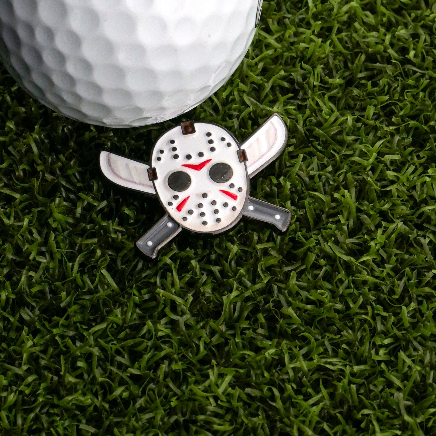 The Mask & Machetes Ball Marker by Ballmarkers.com, shaped like a hockey mask with crossed machetes and golf clubs, rests on artificial grass—an edgy accessory for bold golfers.