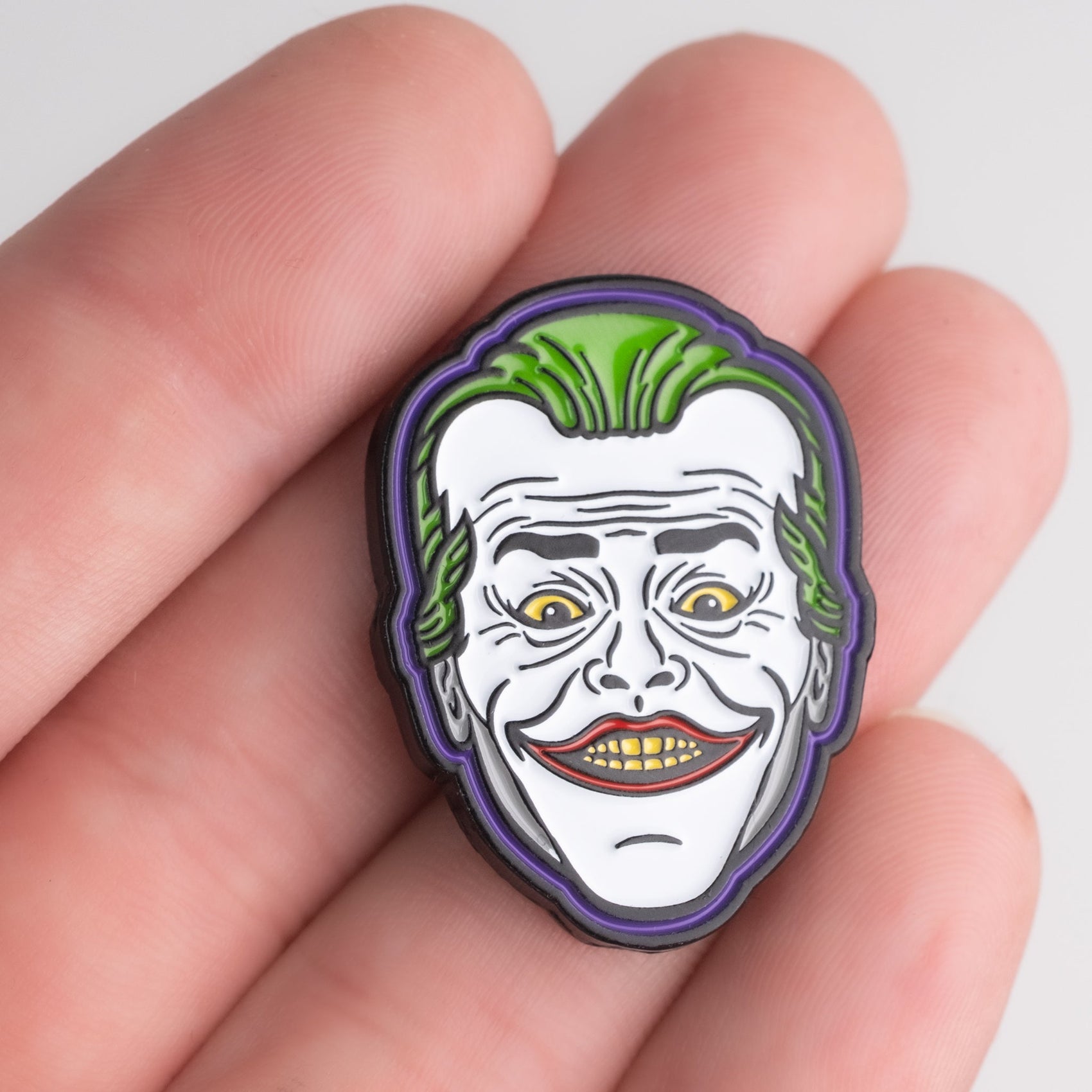 A hand holds the Pins and Aces Joker Ball Marker 3.0, a small enamel golf marker pin featuring a grinning face with green hair, yellow teeth, and comic book villain-style features.
