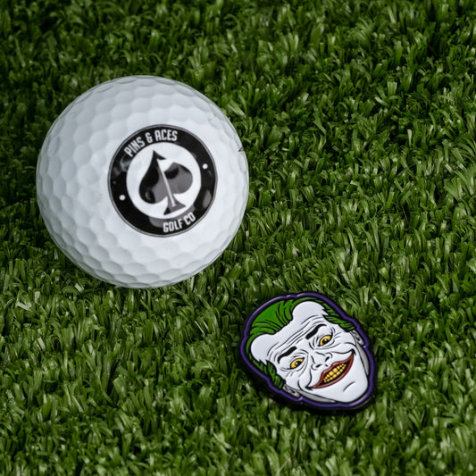 A Pins and Aces golf ball rests on green grass beside the Joker Ball Marker 3.0, which displays a cartoon face with green hair and a wide, sinister smile.