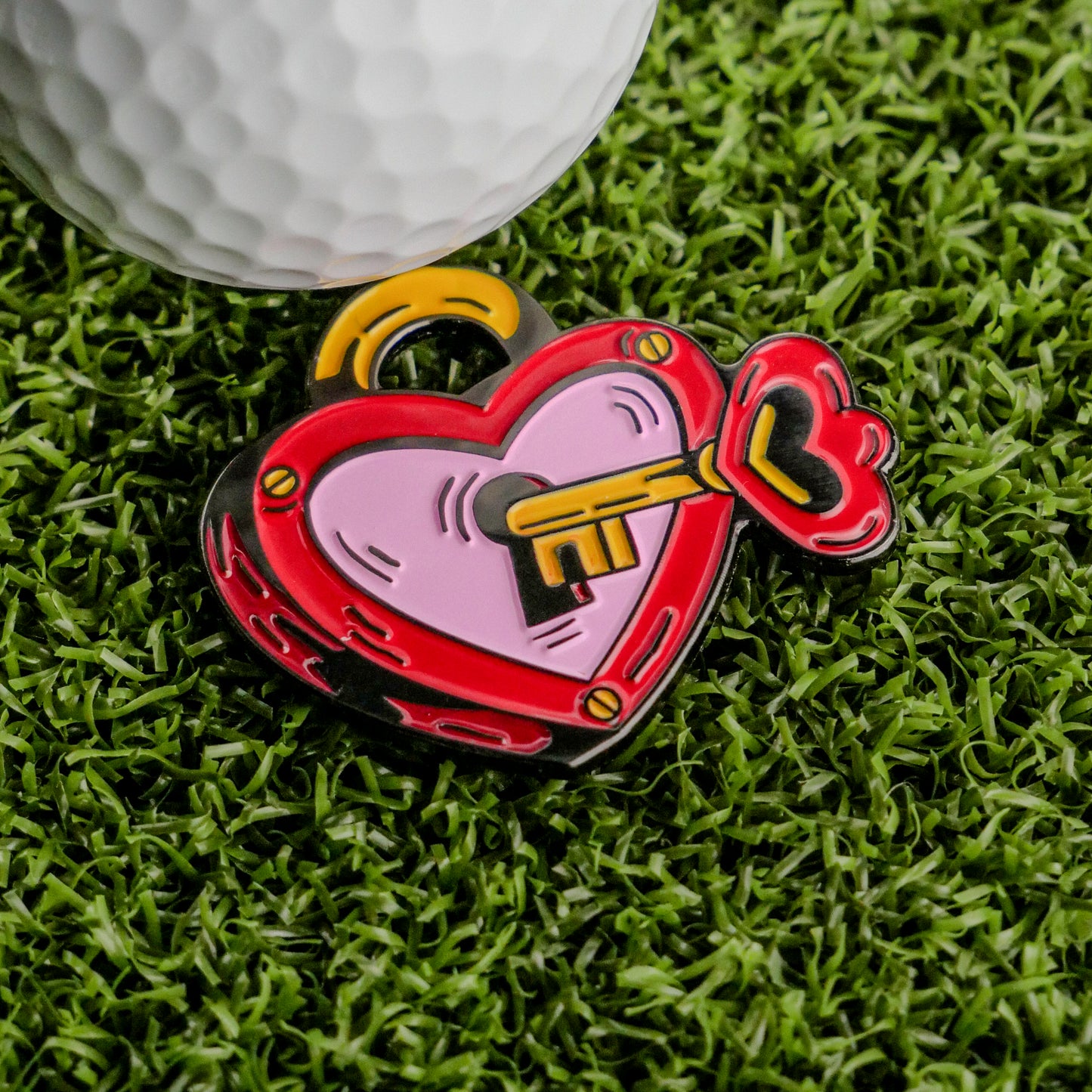 The Key to Your Heart Ball Marker by Ballmarkers.com, a vibrant heart-shaped golf accessory, makes the perfect Valentine’s Day gift. It’s pictured on green artificial grass beside a white golf ball.
