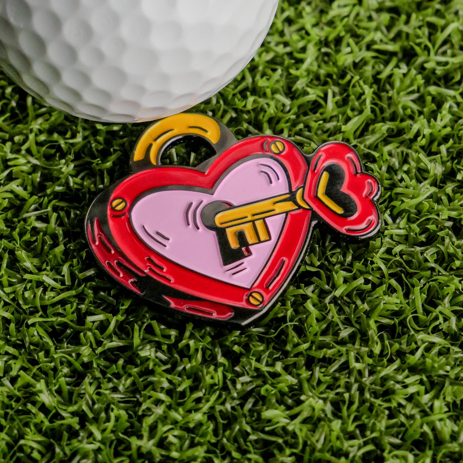 The Key to Your Heart Ball Marker by Ballmarkers.com, a vibrant heart-shaped golf accessory, makes the perfect Valentine’s Day gift. It’s pictured on green artificial grass beside a white golf ball.
