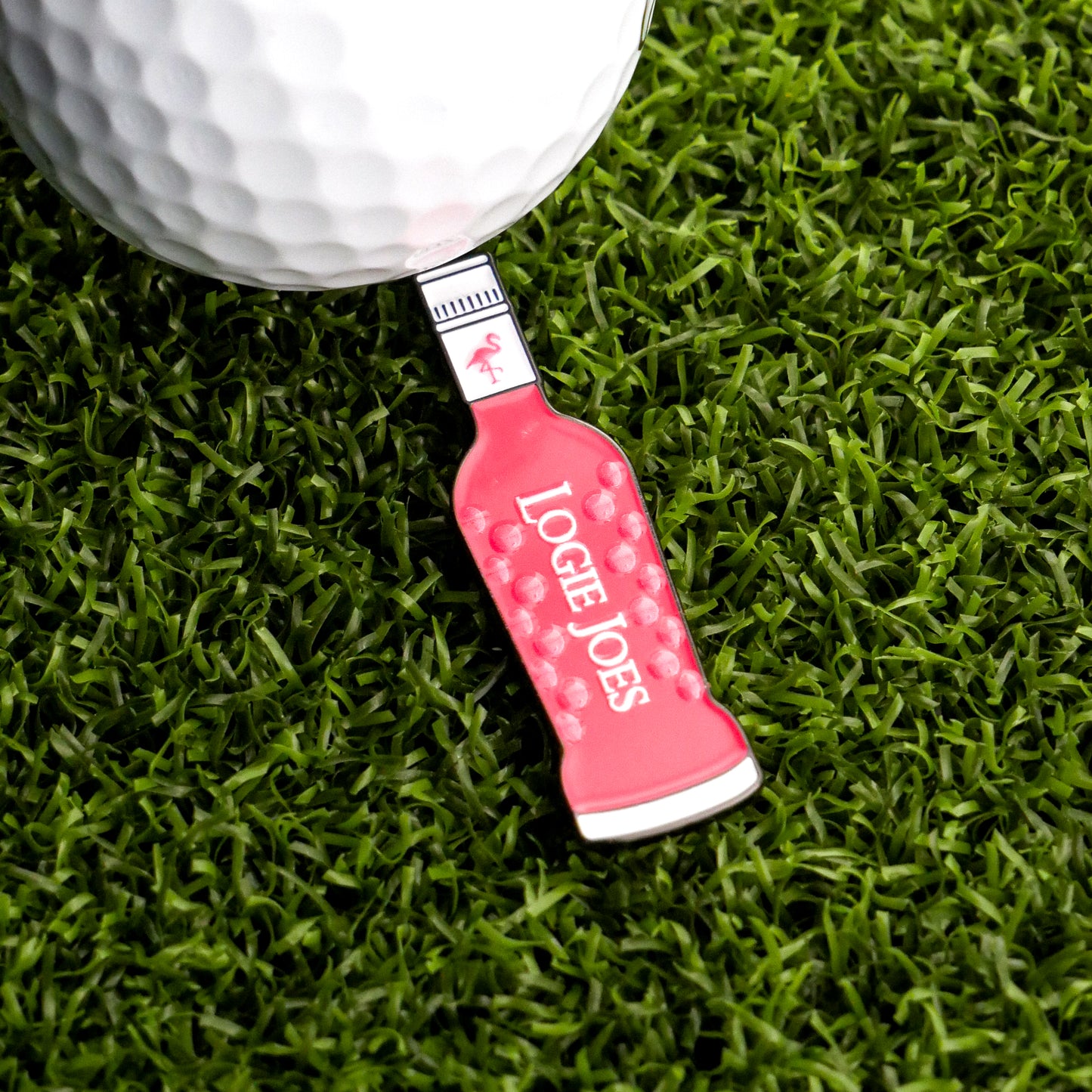 A pink vodka bottle-shaped ball marker from Logie Joe's Golf, labeled "Pink Vodka Ball Marker," sits on artificial green turf next to a white golf ball.