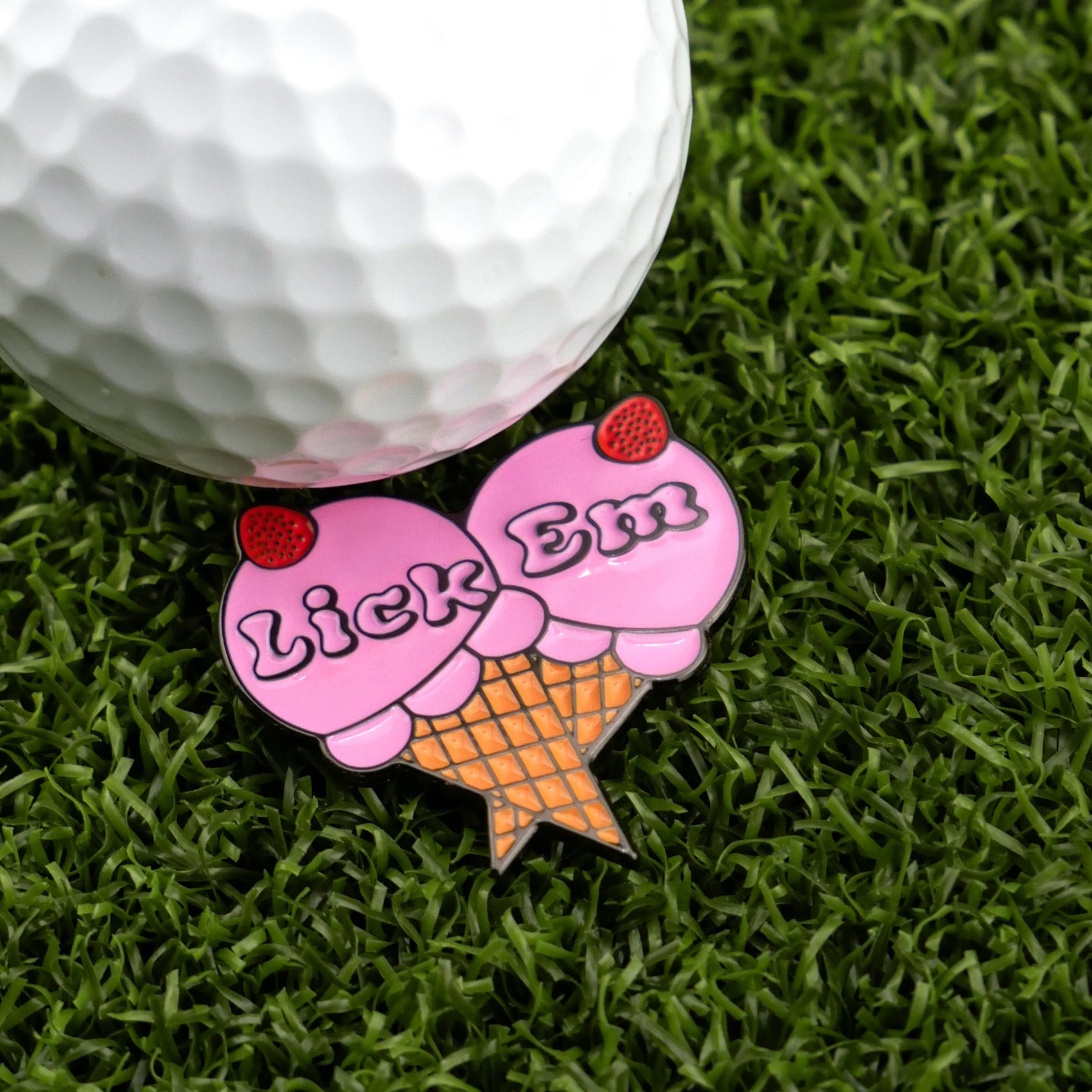 A white golf ball rests on grass beside Logie Joe's Golf Lick Em pin—a unique golf accessory shaped like two pink ice cream scoops in a cone with strawberries, ideal for fun and playful golf ball markers.