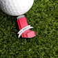The Lil Weezy Ball Marker by Ballmarkers.com, a red inhaler-shaped golf accessory, sits on green grass beside a white golf ball.