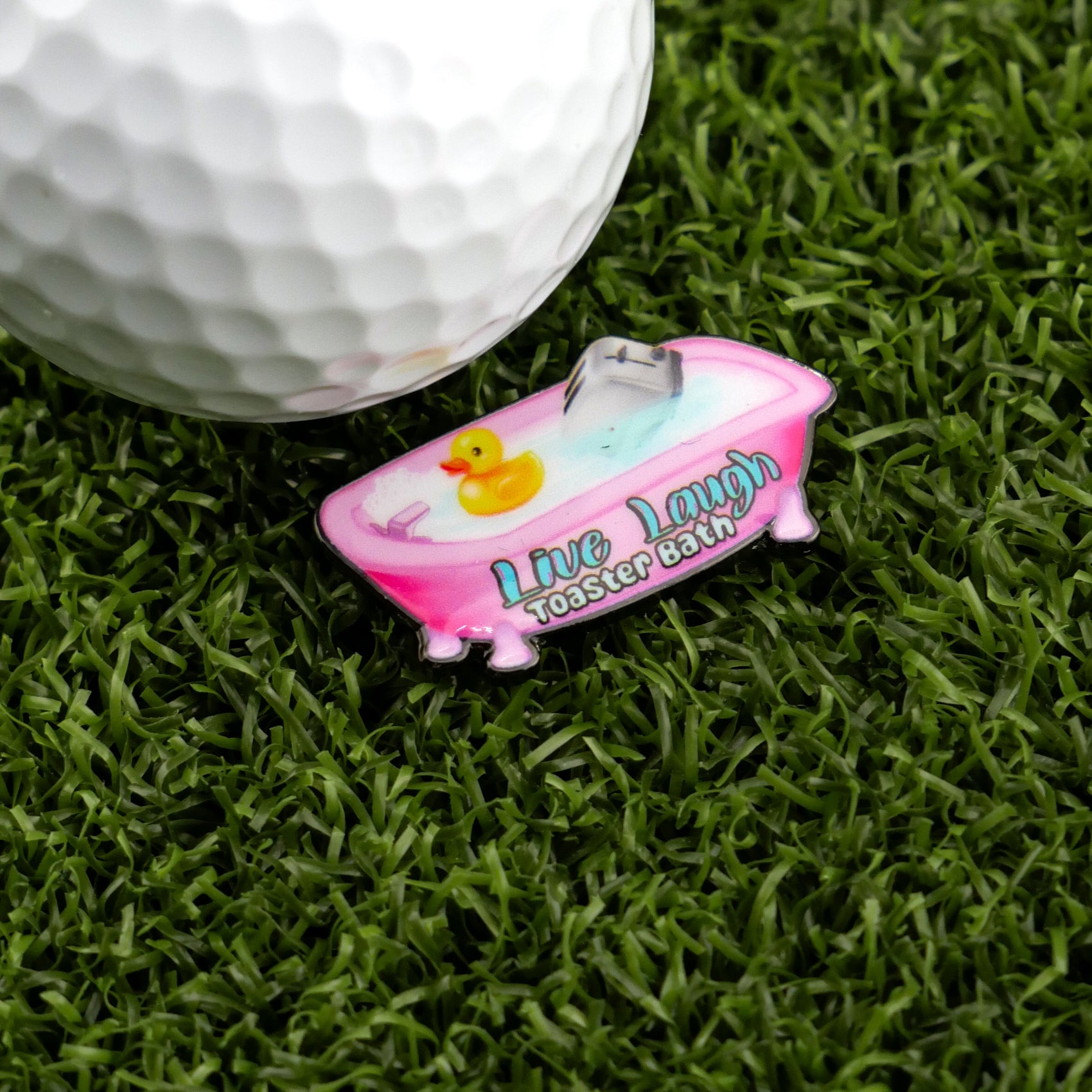 A golf ball on artificial grass beside Logie Joe's Golf Live Laugh Toaster Bath pin—a pink bathtub with a yellow duck—adds a quirky touch to your premium golf accessories or unique golf ball markers.