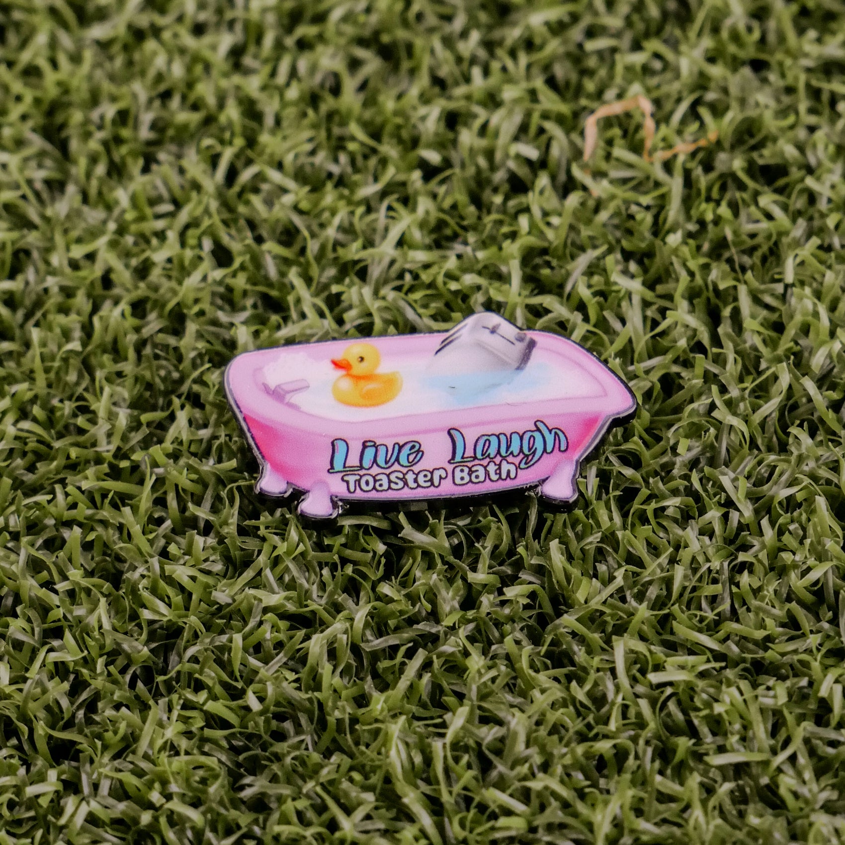 The “Live Laugh Toaster Bath” pin by Logie Joe’s Golf features a pink bathtub with water, a yellow rubber duck, and a toaster on green grass—an offbeat accessory to complement your premium golf ball markers and divot tools.