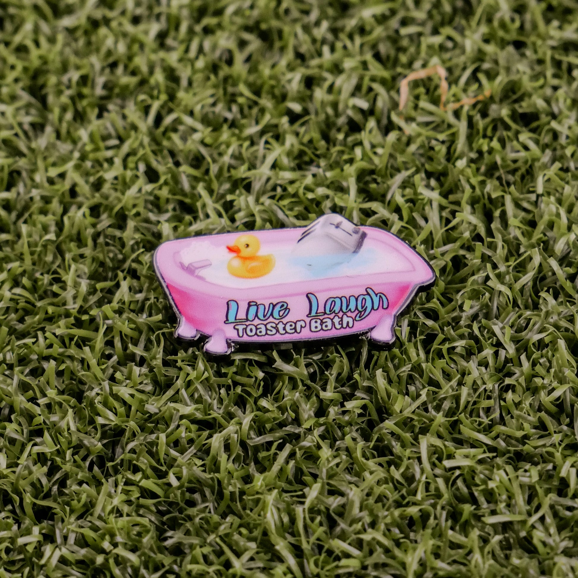 The “Live Laugh Toaster Bath” pin by Logie Joe’s Golf features a pink bathtub with water, a yellow rubber duck, and a toaster on green grass—an offbeat accessory to complement your premium golf ball markers and divot tools.