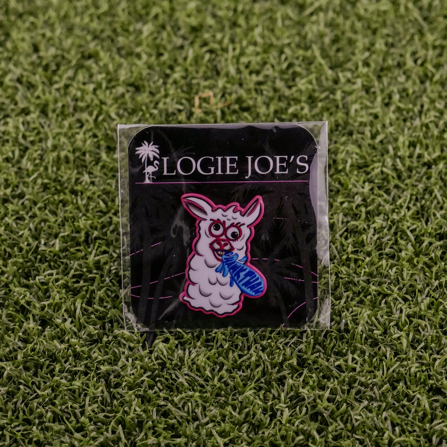 The Hawk Tuah Llama pin by Logie Joe's Golf features a cartoon llama with a blue fish, displayed on a black LOGIE JOE'S card atop green turf—perfect for fans of unique golf accessories or playful ball markers.