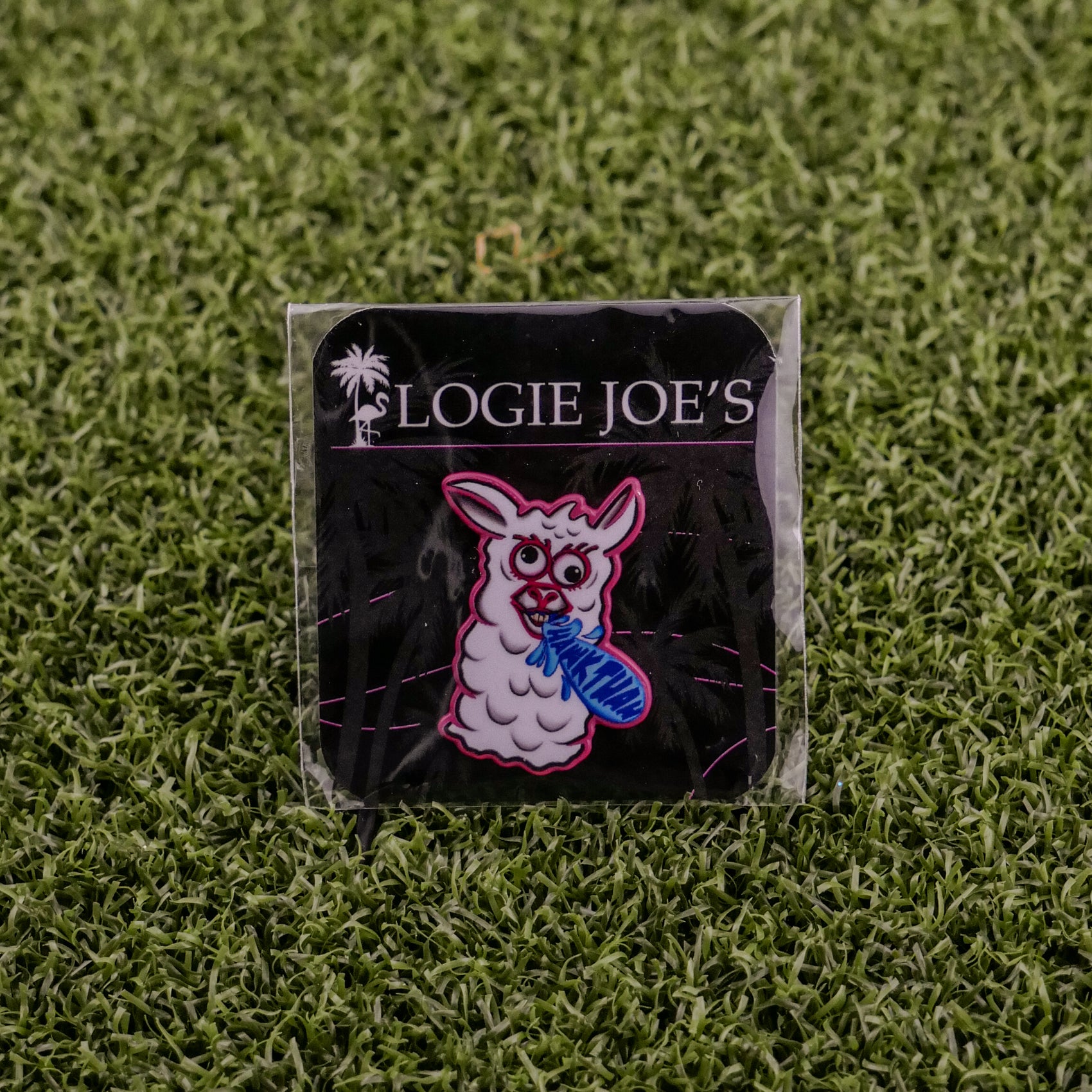 The Hawk Tuah Llama pin by Logie Joe's Golf features a cartoon llama with a blue fish, displayed on a black LOGIE JOE'S card atop green turf—perfect for fans of unique golf accessories or playful ball markers.
