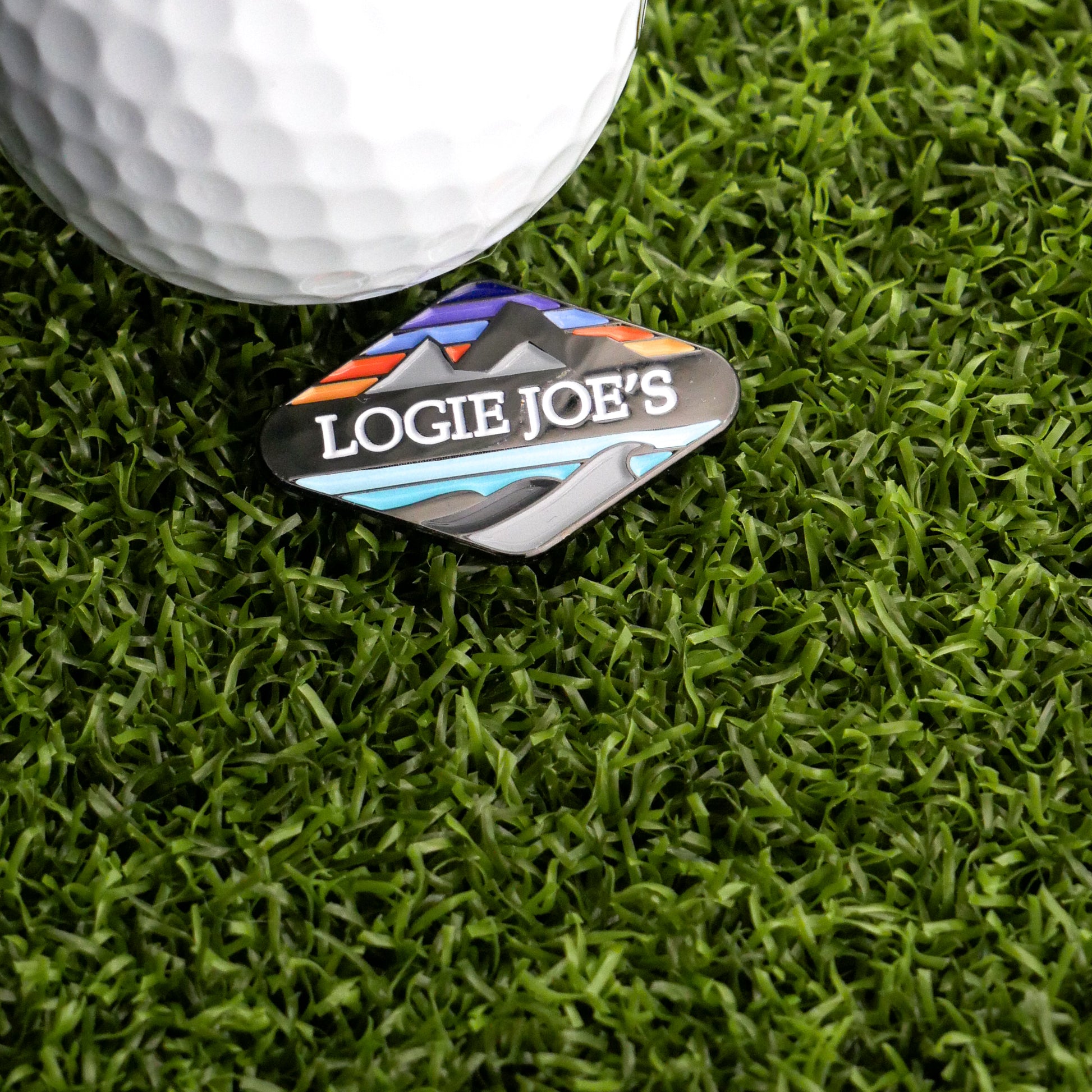 A golf ball on artificial grass sits next to the Logie Joe's Golf Ball Marker, featuring the Logie Joe's Golf mountain wave logo and colorful landscape artwork.