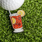 A golf ball rests on green grass beside the Ballmarkers.com Long Island Iced Tea Ball Marker—a unique addition to any golfer’s cocktail-themed accessory collection.