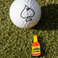 A golf ball with a black spade symbol rests on green grass beside the Pins and Aces Louisiana Hot Sauce Ball Marker, shaped like a small yellow sauce bottle.
