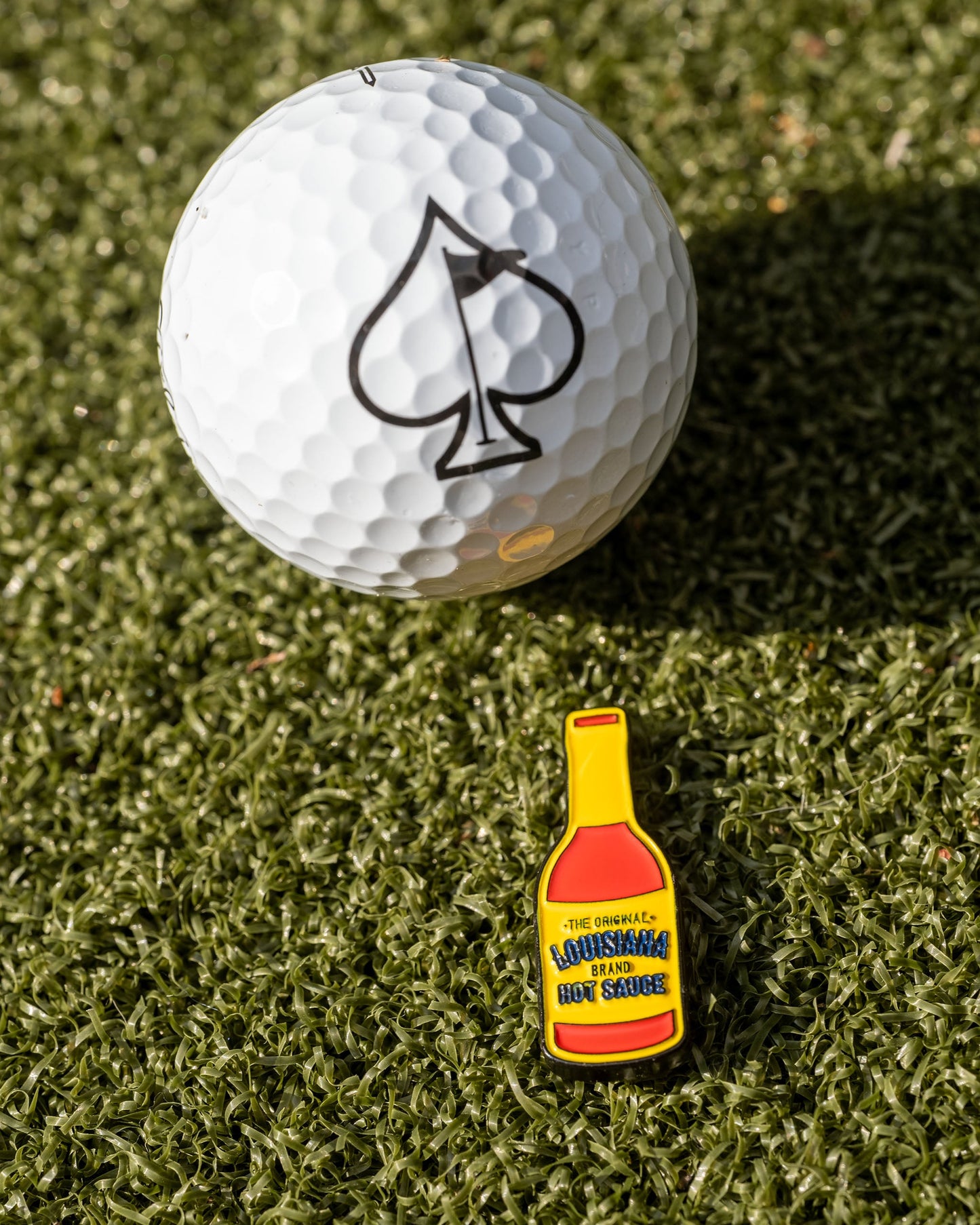 A golf ball with a black spade symbol rests on green grass beside the Pins and Aces Louisiana Hot Sauce Ball Marker, shaped like a small yellow sauce bottle.