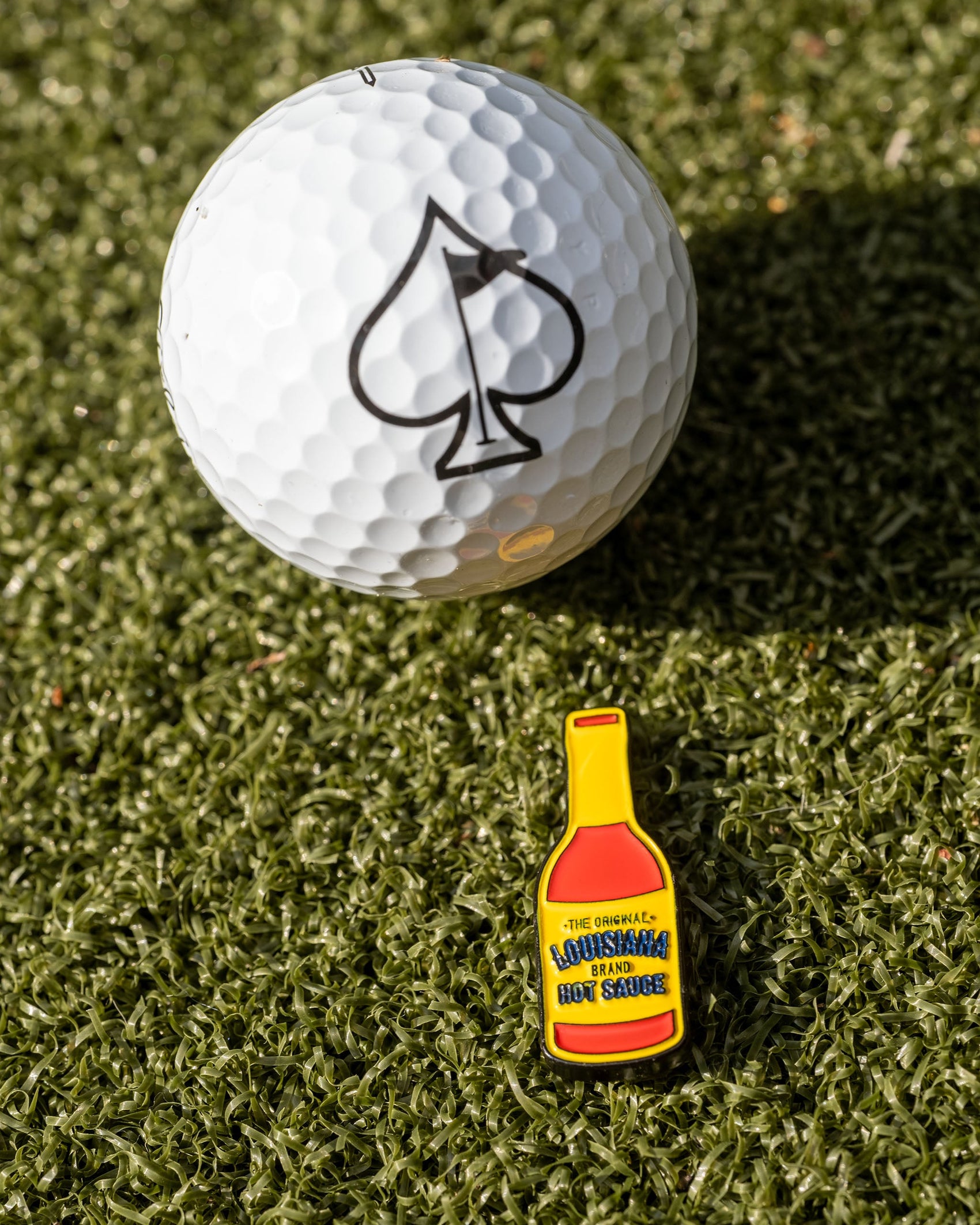 A golf ball with a black spade symbol rests on green grass beside the Pins and Aces Louisiana Hot Sauce Ball Marker, shaped like a small yellow sauce bottle.