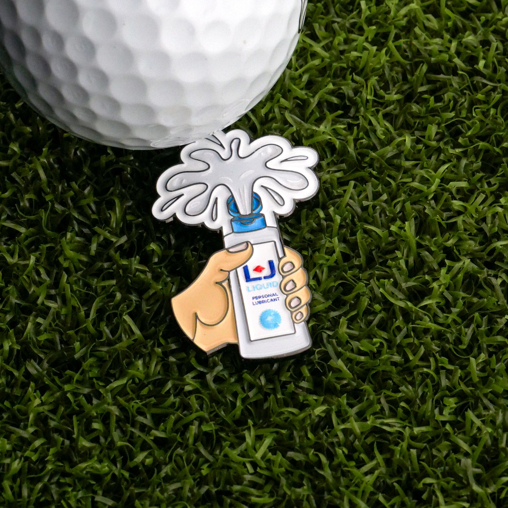 The Logie Joe's Golf Personal Lubricant Ball Marker is a decorative pin shaped like a hand with a spray bottle, shown on artificial grass next to a low-friction golf ball marker designed for maximum speed and roll.
