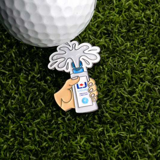 The Logie Joe's Golf Personal Lubricant Ball Marker is a decorative pin shaped like a hand with a spray bottle, shown on artificial grass next to a low-friction golf ball marker designed for maximum speed and roll.