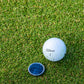 A white golf ball and a blue round Carbon Fiber "The Venom" ball marker by Doubloon rest on the green grass of a golf course.