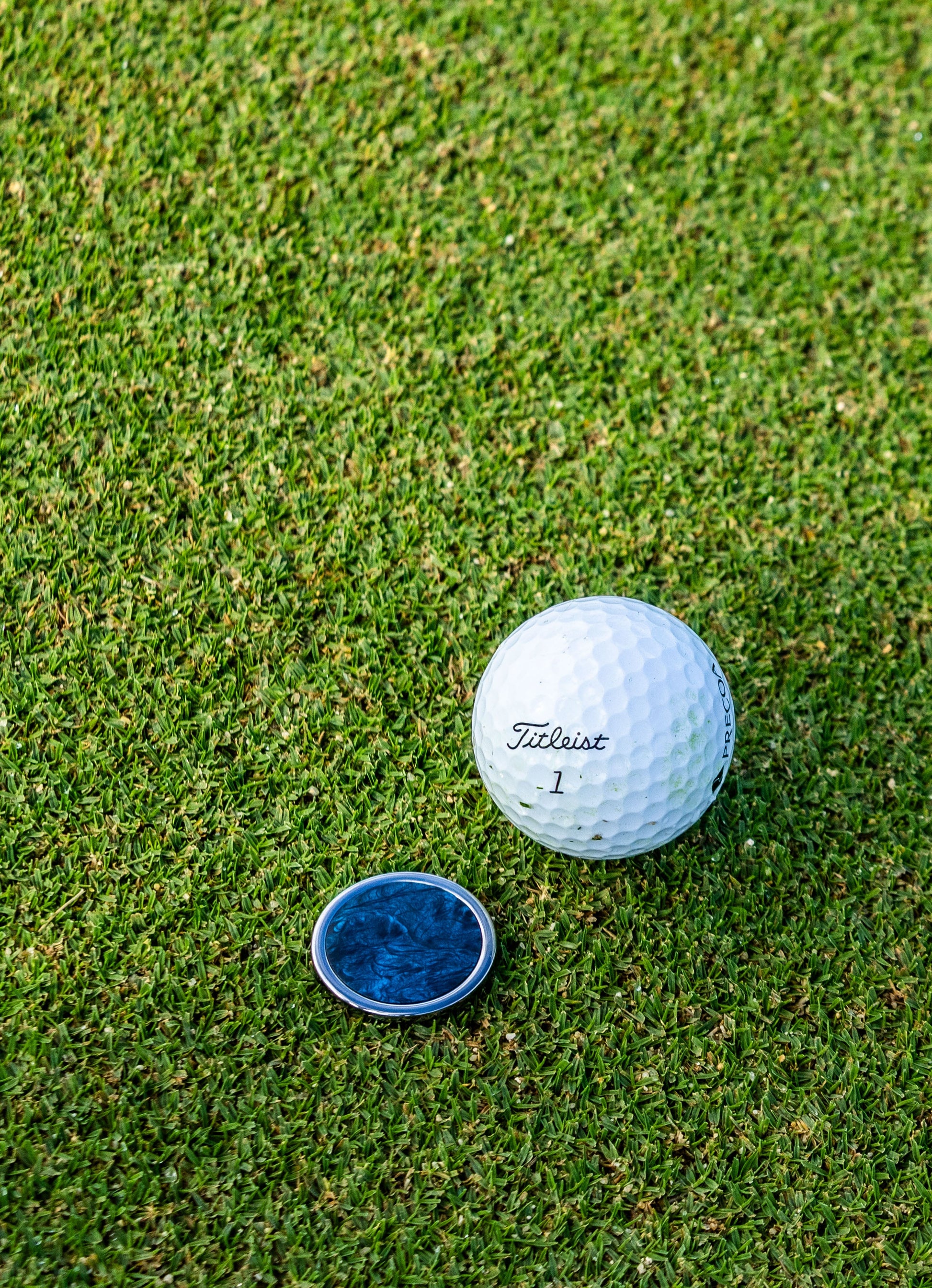 A white golf ball and a blue round Carbon Fiber "The Venom" ball marker by Doubloon rest on the green grass of a golf course.