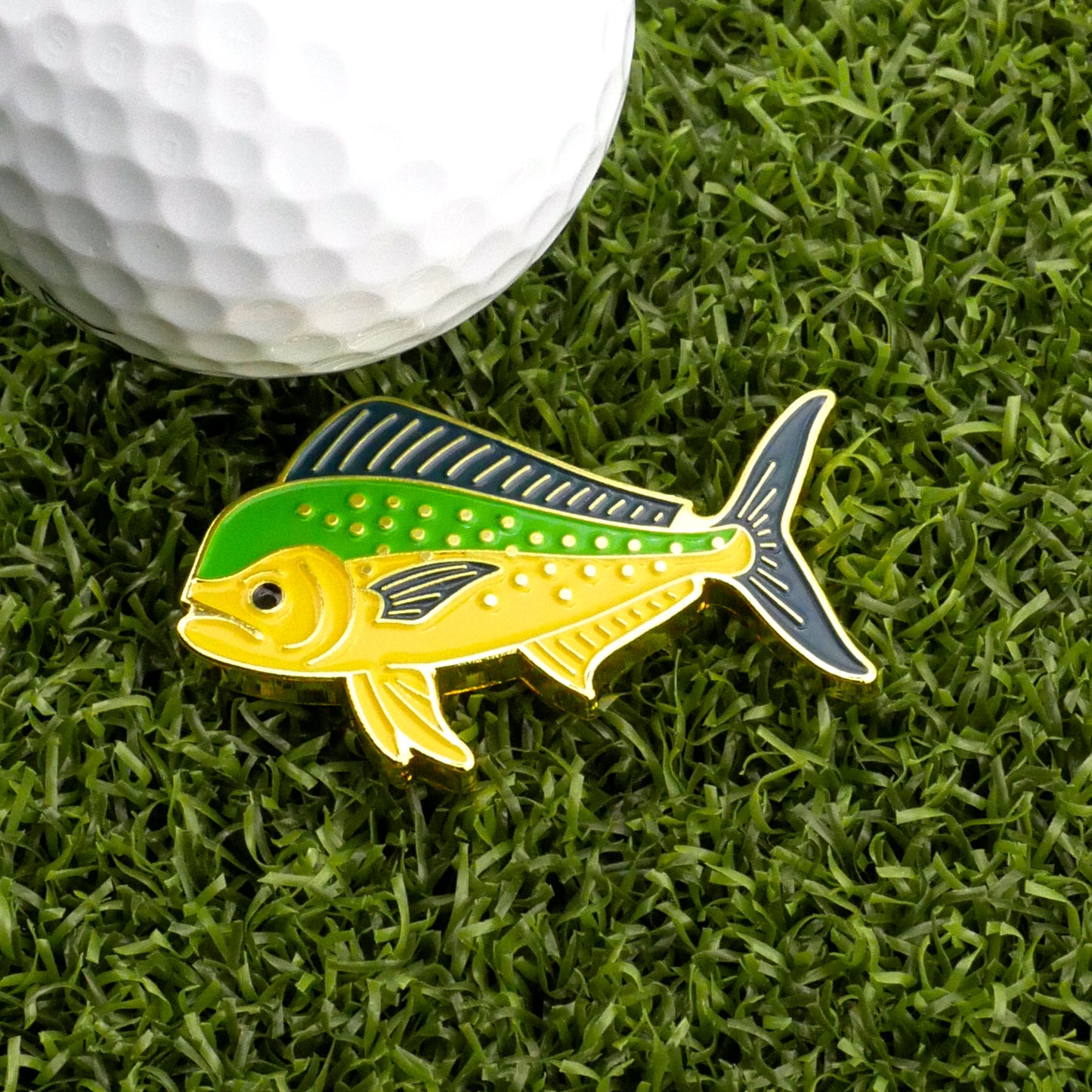 The Logie Joe's Golf Mahi Mahi Ball Marker, featuring green and yellow accents, rests on artificial grass beside a white golf ball.