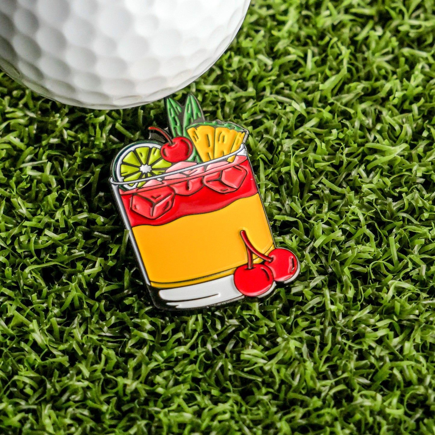 The Mai Tai Ball Marker from Ballmarkers.com—a vibrant enamel pin shaped like a tropical cocktail with fruit and ice—rests on green turf by a white golf ball, perfect for your collection of unique golf accessories.