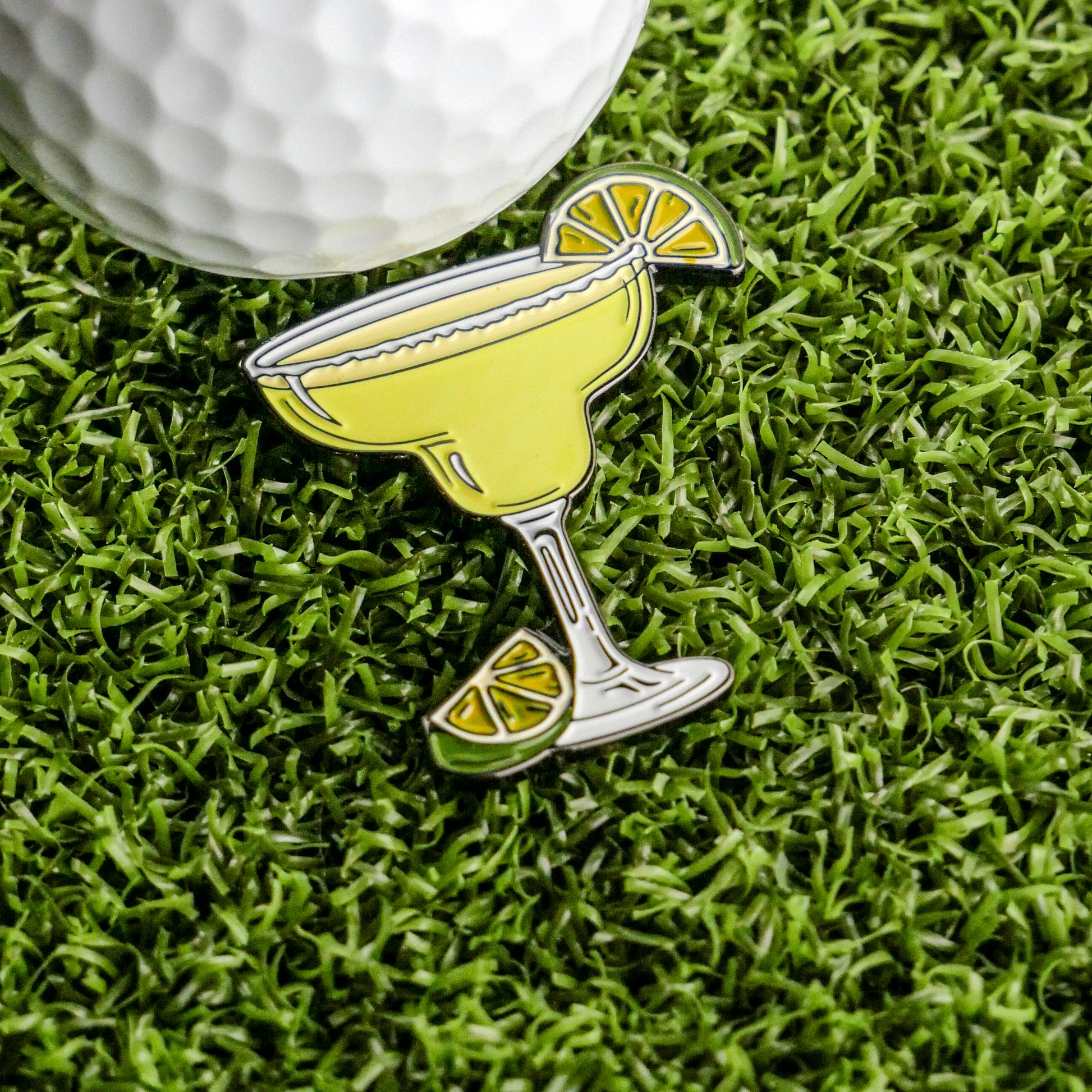 The Margarita Ball Marker by Ballmarkers.com features a detailed enamel pin with a lemon slice, shown on artificial green grass beside a white golf ball—an ideal fun accessory for your golf collection.