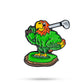 The Margaritaville - Golfing Parrot Ball Marker by Pins and Aces features a colorful green parrot with an orange chest and yellow beak, standing on a small green platform, golf club over its shoulder, ready to swing.