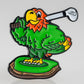 A colorful cartoon parrot with green feathers and an orange head stands on grass, holding a golf club and smiling—featured on the Margaritaville - Golfing Parrot Ball Marker by Pins and Aces.