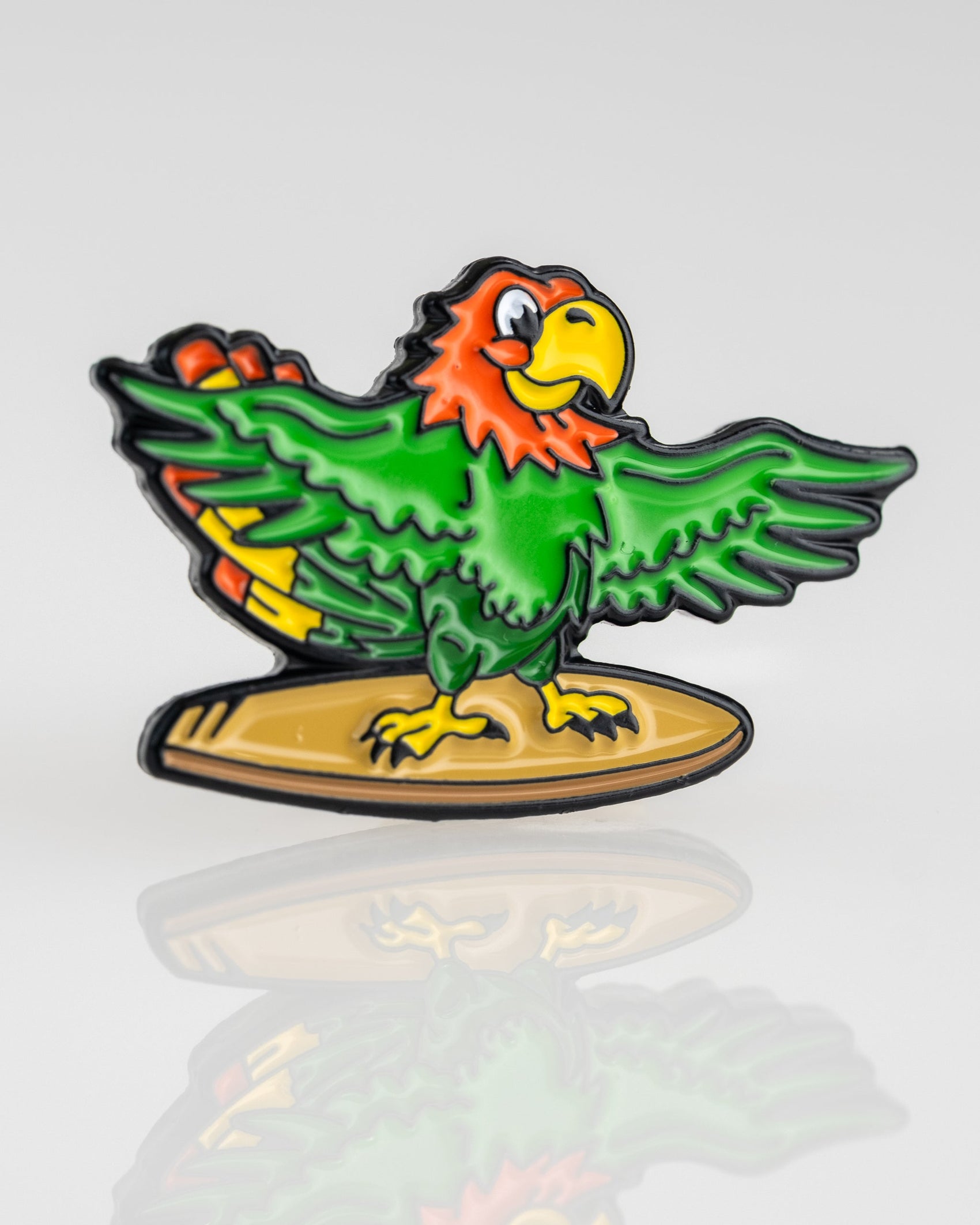 A colorful cartoon parrot with red, green, and yellow feathers stands on a yellow surfboard, wings spread wide. Inspired by Pins and Aces, this Margaritaville - Surfing Parrot Ball Marker is shown against a plain white background.