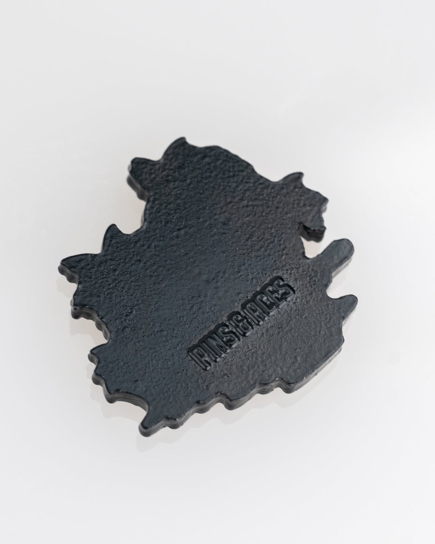 A black, irregularly shaped Margaritaville - Sleeping Parrot Ball Marker by Pins and Aces rests on a white surface, showcasing the brand's signature style.