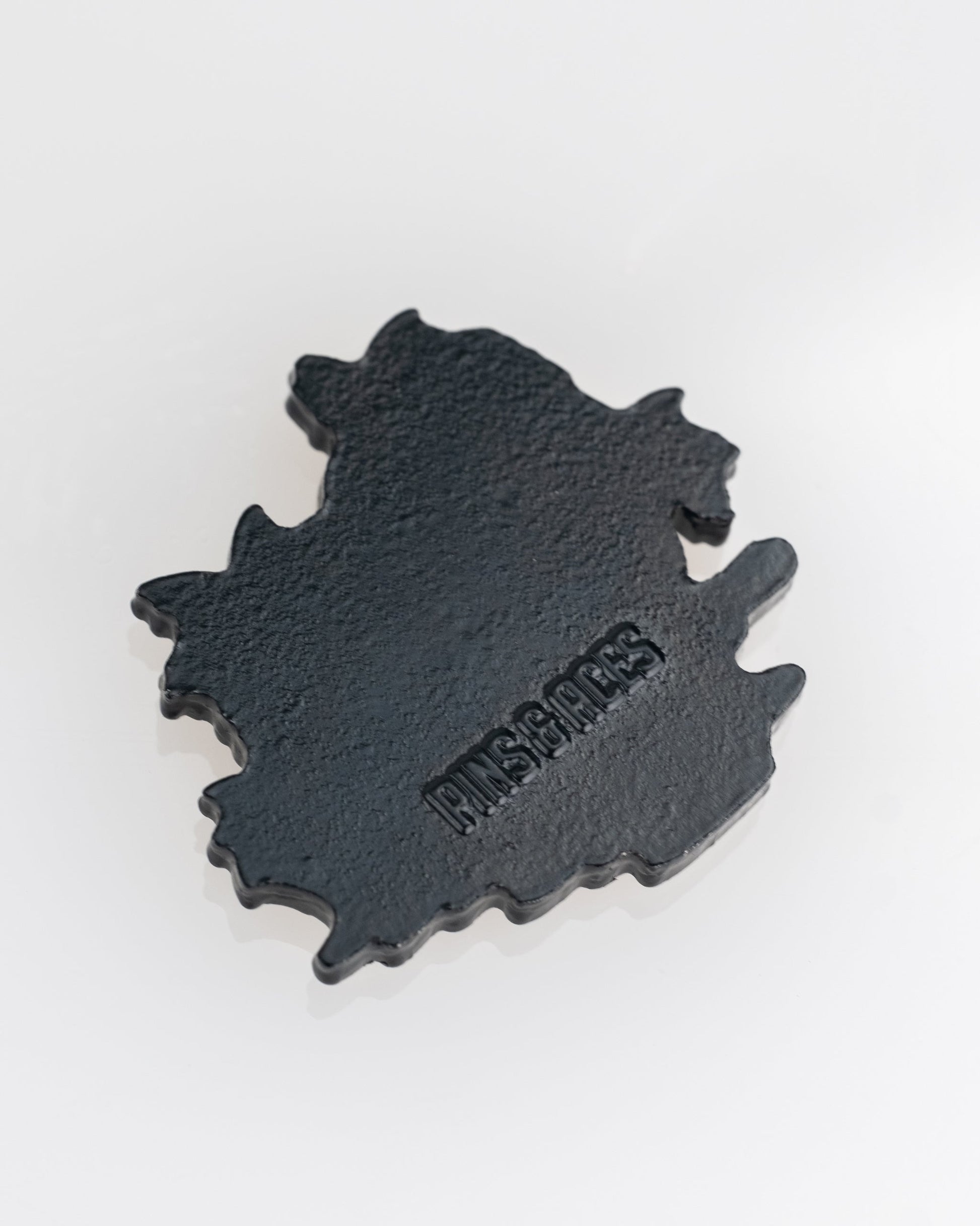 A black, irregularly shaped Margaritaville - Sleeping Parrot Ball Marker by Pins and Aces rests on a white surface, showcasing the brand's signature style.