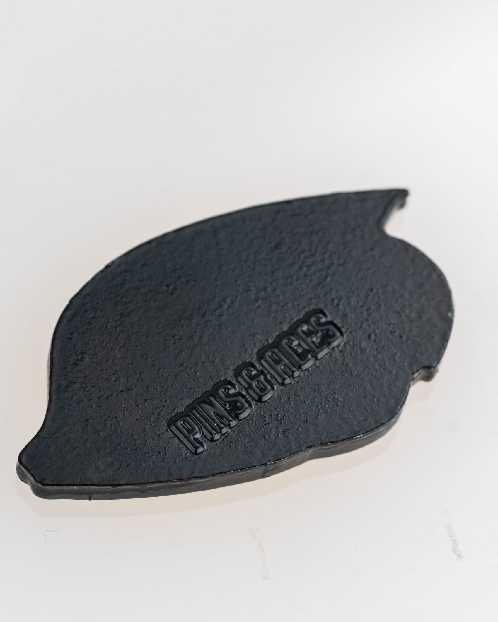 A black, leaf-shaped Margaritaville - Angel Fish Ball Marker by Pins and Aces, featuring a rough texture and embossed branding, is shown on a light, reflective background.