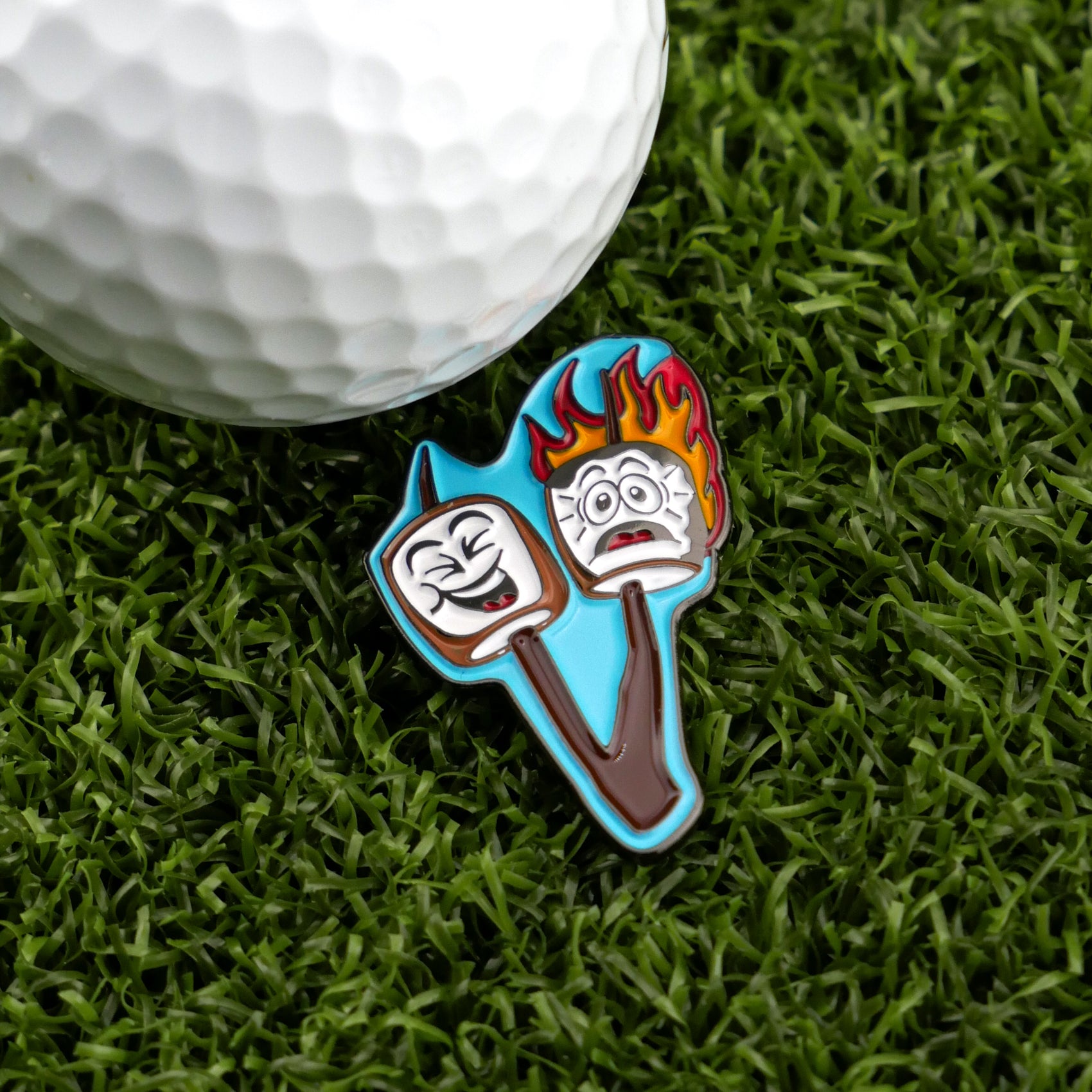 A golf ball on grass sits beside Logie Joe's Golf "Marshmallows" ball marker, featuring two cartoon marshmallows—one smiling, one scared and aflame—for a hilarious golf accessory.