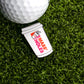 A Logie Joe's Golf Mass Holes Coffee Ball Marker, shaped like a coffee cup with bold red and orange lettering, sits next to a golf ball on artificial grass.