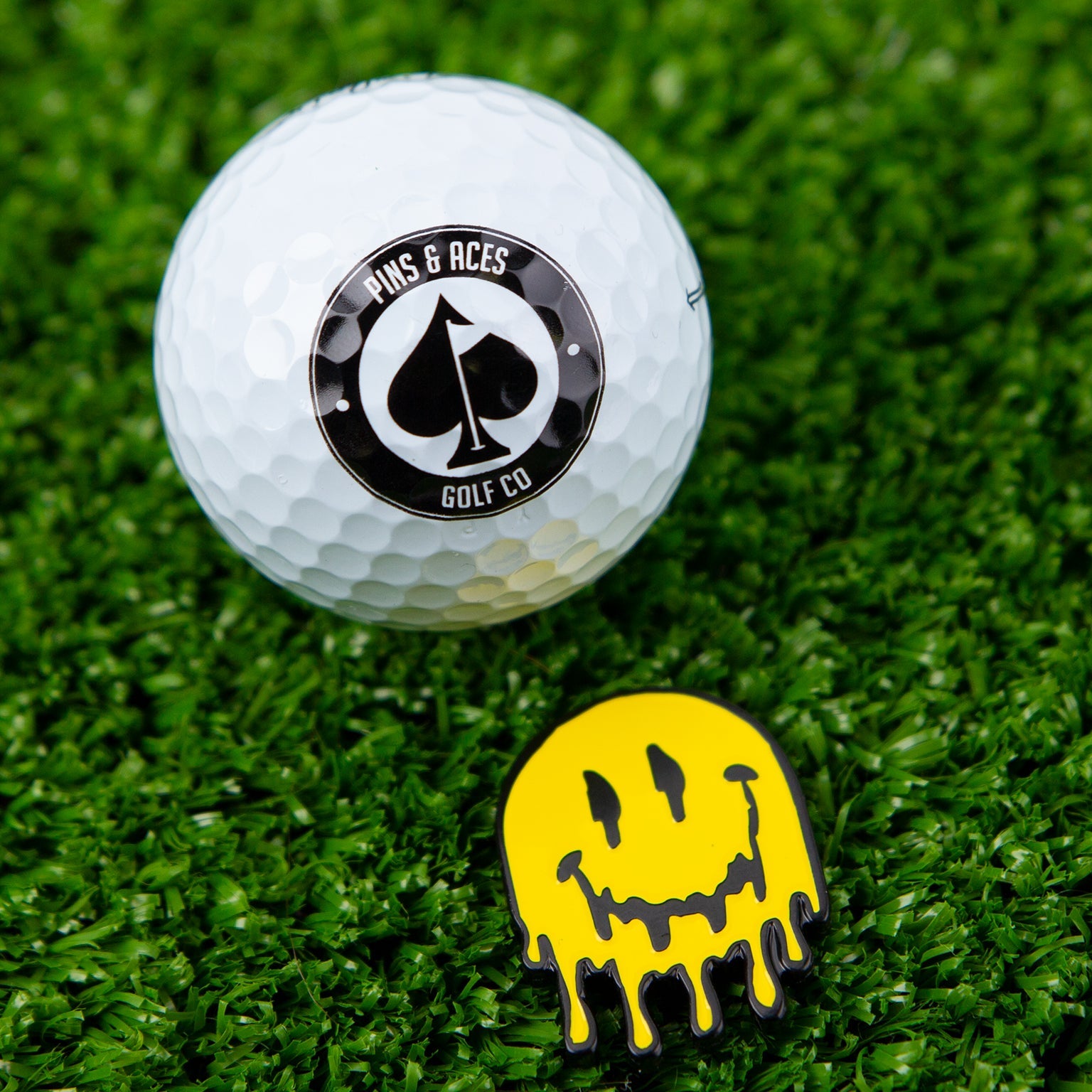 A white golf ball with a Pins and Aces logo sits beside the Melting Face Ball Marker—a yellow, dripping smiley face marker made of stainless steel. Both items rest on green grass.