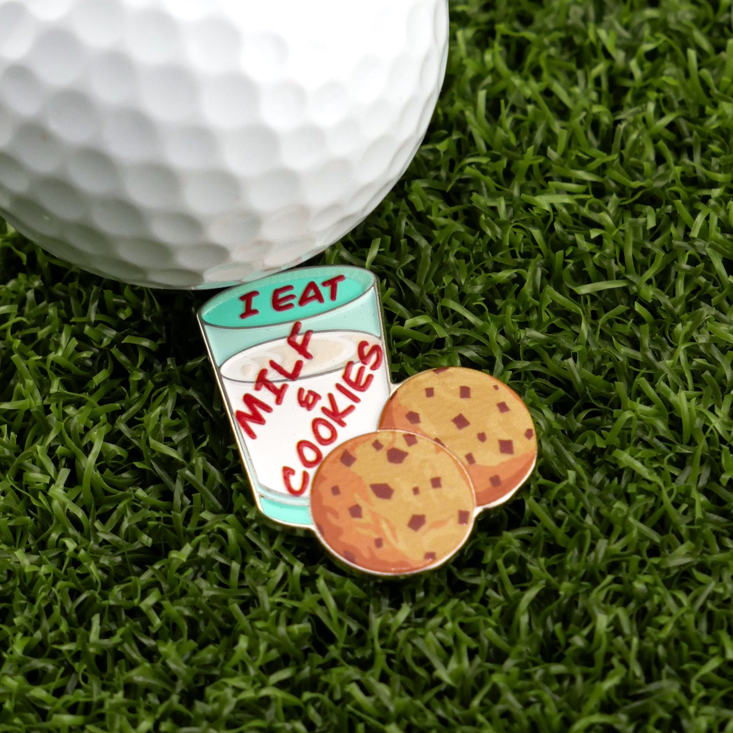 The Logie Joe's Golf "Milf & Cookies" golf ball marker features a milk glass-shaped pin labeled “I EAT MILK & COOKIES,” two chocolate chip cookies, and a premium golf ball, making it a fun addition to any collection.