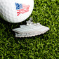A golf ball with an American flag design sits on grass next to the silver Aircraft Carrier Ball Marker by Ballmarkers.com, honoring military aviation.