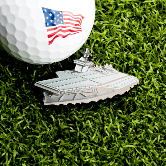 A golf ball with an American flag design sits on grass next to the silver Aircraft Carrier Ball Marker by Ballmarkers.com, honoring military aviation.