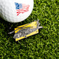 A golf ball with an American flag design rests on green grass beside the Ballmarkers.com Apache Helicopter Ball Marker, a distinctive yellow-background golf accessory.