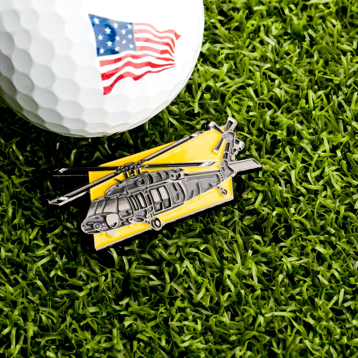 A golf ball with an American flag design rests on green grass beside the Ballmarkers.com Apache Helicopter Ball Marker, a distinctive yellow-background golf accessory.