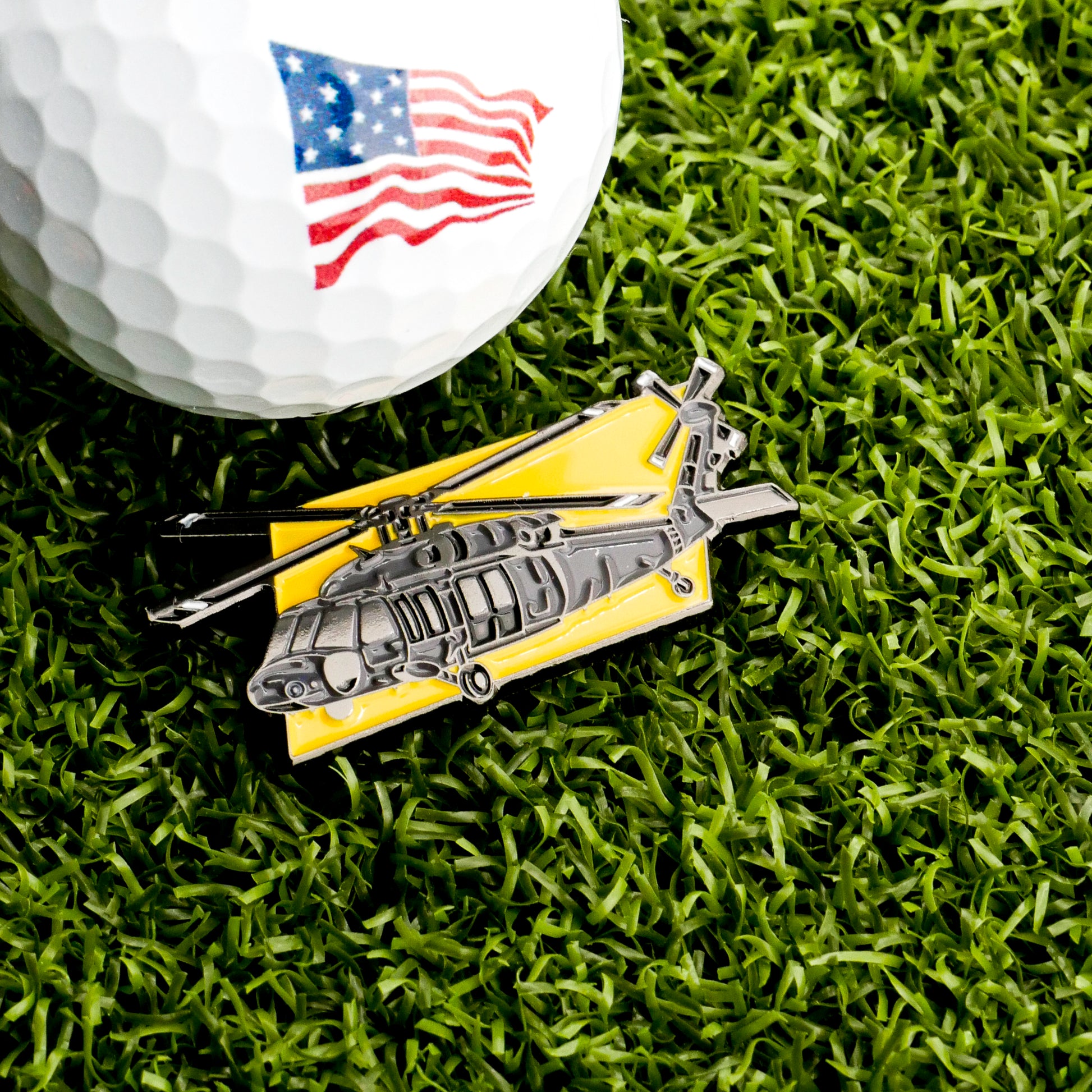 A golf ball with an American flag design rests on green grass beside the Ballmarkers.com Apache Helicopter Ball Marker, a distinctive yellow-background golf accessory.