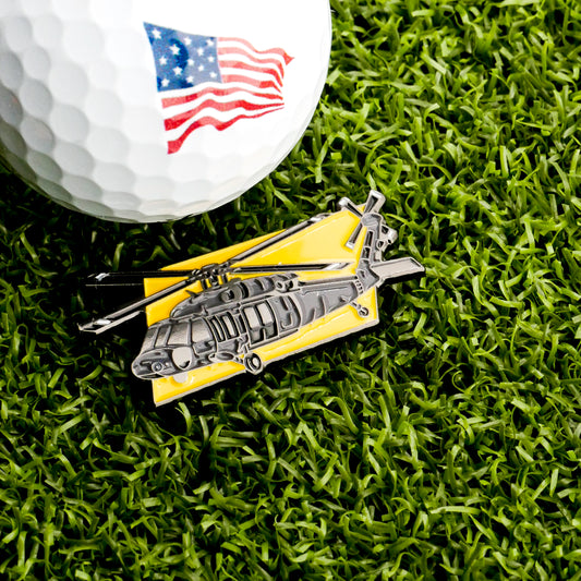 A golf ball with an American flag design rests on green grass beside the Ballmarkers.com Apache Helicopter Ball Marker, a distinctive yellow-background golf accessory.