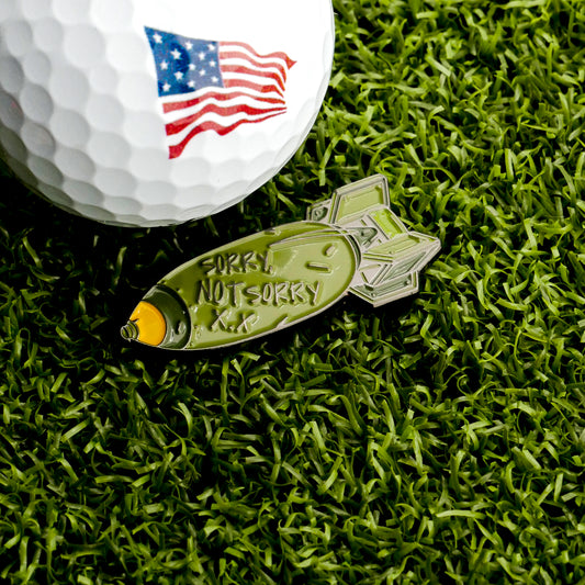 A golf ball with an American flag graphic rests on grass beside the Sorry, Not Sorry Bomb Ball Marker from Ballmarkers.com—a metal pin-shaped marker engraved with “SORRY NOT SORRY” for a unique military-themed golf accessory.