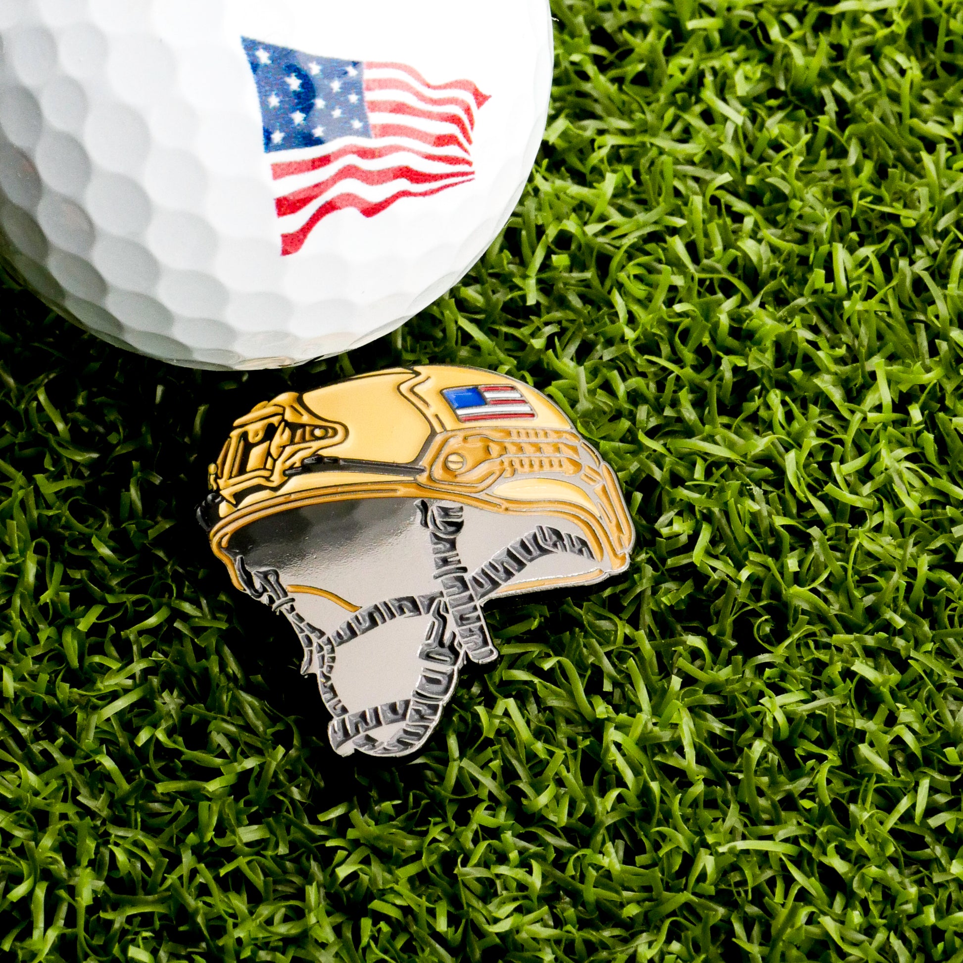 A close-up of a golf ball with an American flag design and the Ballmarkers.com Combat Helmet Ball Marker, both displayed on green artificial grass, showcases this unique military-themed golf accessory.