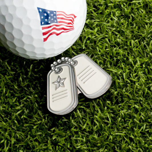 The Ballmarkers.com Dog Tags Ball Marker, featuring a patriotic American flag design, sits on green grass—an ideal golf accessory for those who want a military-inspired touch on the course.