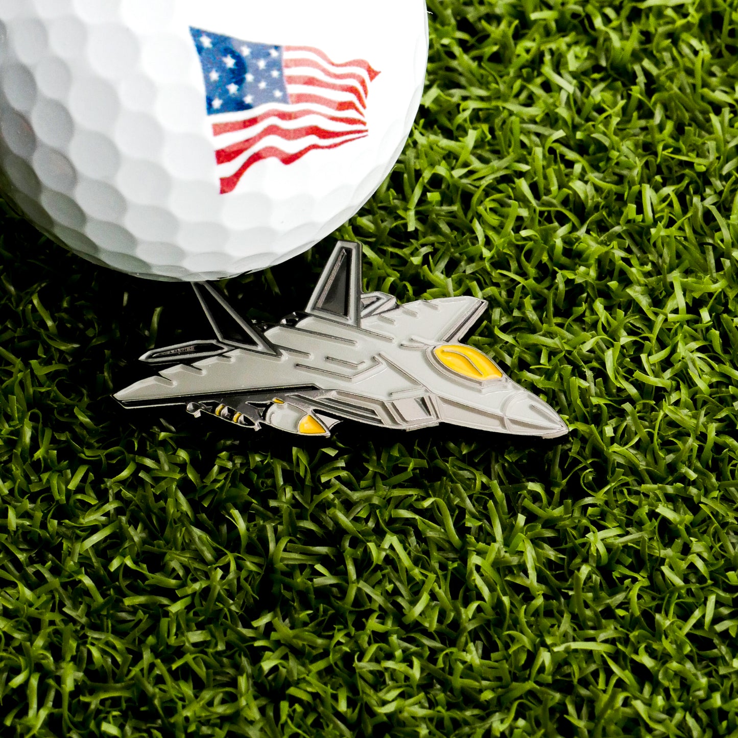 A golf ball with an American flag design sits on artificial grass beside the Fighter Jet Ball Marker by Ballmarkers.com—the perfect aviation golf accessory for enthusiasts.